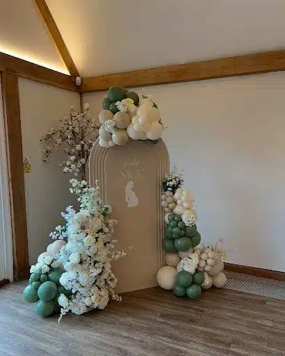 Elegant Arch with Organic Balloon Garland and Floral Accents
This charming baby shower backdrop exudes a sense of natural elegance. A soft beige arched panel serves as the focal point, delicately inscribed with "Baby NICE" and featuring a subtle silhouette of a pregnant woman. The arch is beautifully adorned with an organic balloon garland in varying shades of sage green and cream, creating a visually appealing and modern aesthetic.

Adding to the enchantment are lush floral arrangements cascading from both sides of the arch. White roses and delicate blossoms, interspersed with greenery, bring a touch of freshness and sophistication to the setup. The asymmetrical placement of the balloons and flowers contributes to a relaxed yet stylish ambiance, perfect for celebrating the upcoming arrival.

This backdrop idea beautifully blends contemporary design with classic floral elements, offering a sophisticated and memorable setting for baby shower photos and celebrations. The neutral color palette allows it to seamlessly integrate with various party themes and color schemes, making it a versatile and chic choice.