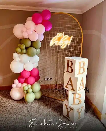 
Playful Neon Sign with Balloon Garland and Letter Blocks
This vibrant baby shower backdrop features a playful combination of modern and classic elements. A golden wire mesh arch serves as the frame, showcasing a bright neon sign that spells out "Oh baby!". The warm glow of the neon adds a fun and celebratory touch to the setting.

An organic balloon garland, cascading down the side of the arch, introduces a burst of color and whimsy. The garland incorporates a mix of pink, white, and sage green balloons, creating a visually appealing and dynamic arrangement. The varying sizes and shades of the balloons contribute to a relaxed yet stylish ambiance.

Adding a touch of nostalgia and charm are large white letter blocks, spelling out "BABY" with golden lettering. These blocks provide a delightful focal point and enhance the celebratory atmosphere. This backdrop idea beautifully blends contemporary design with classic elements, offering a memorable and photogenic setting for a baby shower.